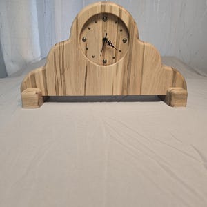 May include: A wooden clock with a light brown finish and a white face. The clock has a unique, cloud-like shape and is made of wood with a natural grain pattern.