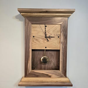 May include: A wooden wall clock with a pendulum and a brass weight. The clock face is made of light wood and has black hands. The frame is made of dark wood with a light wood trim.