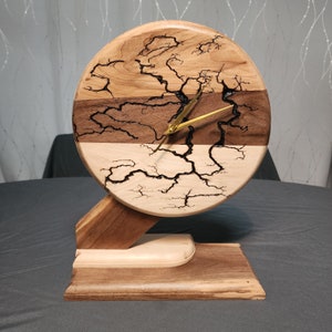 Op de afbeelding: Een houten klok met een ronde wijzerplaat en een uniek ontwerp. De wijzerplaat is gemaakt van drie verschillende soorten hout, elk met een ander nerfpatroon. De wijzers van de klok zijn goudkleurig. De klok staat op een houten voet.