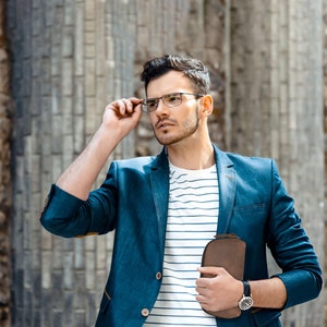Puede incluir: Un hombre con un blazer azul sobre una camisa blanca a rayas, sosteniendo una cartera de cuero marr&oacute;n en su mano derecha. Lleva gafas y tiene barba.