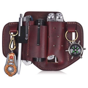 May include: A brown leather utility belt with multiple pockets and loops for carrying tools and accessories. The belt includes a compass, a flashlight, a multi-tool, a pen, and a key chain with a remote control.