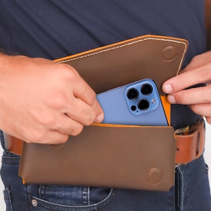 K&ouml;nnte beinhalten: Eine braune Leder-Handytasche mit gelben N&auml;hten. Ein blaues Smartphone mit einer Kamera-Linse ist teilweise in der Tasche sichtbar.