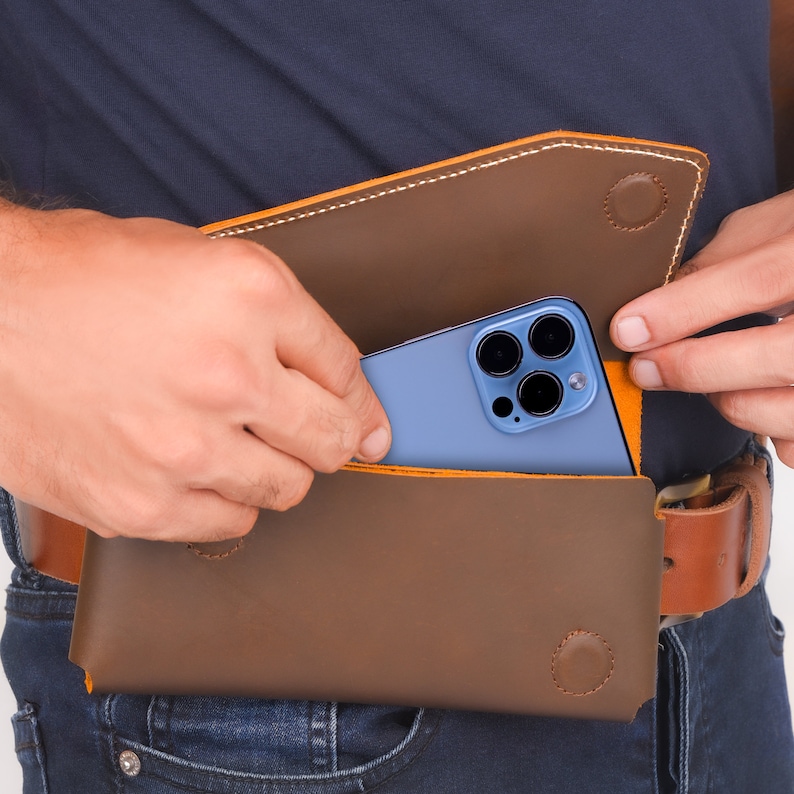 K&ouml;nnte beinhalten: Eine braune Leder-Handytasche mit gelben N&auml;hten. Ein blaues Smartphone mit einer Kamera-Linse ist teilweise in der Tasche sichtbar.