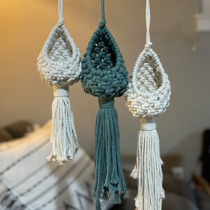 May include: Three macrame plant hangers with a teardrop shape and tassel fringe. The hangers are made with natural cotton rope and are available in white and green.