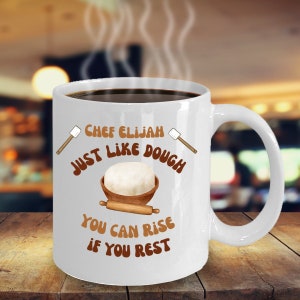 May include: White ceramic coffee mug with a brown design. The mug has the text "Chef Elijah Just Like Dough You Can Rise If You Rest" on it. The design includes a bowl of dough, a rolling pin, and two wooden spatulas.
