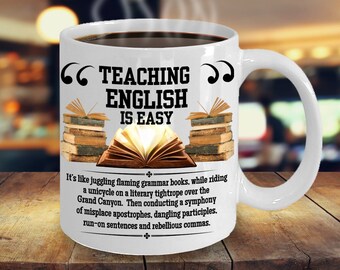 May include: White ceramic coffee mug with a black quote around the text "TEACHING ENGLISH IS EASY". The mug has a black and white illustration of a stack of books and an open book with a yellow glow. The text below the illustration reads "It's like juggling flaming grammar books, while riding a unicycle on a literary tightrope over the Grand Canyon. Then conducting a symphony of misplace apostrophes, dangling participles, run-on sentences and rebellious commas."