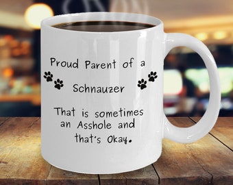 May include: White ceramic coffee mug with black text that reads "Proud Parent of a Schnauzer That is sometimes an Asshole and that's Okay."  Two black paw prints are on either side of the word "Schnauzer".