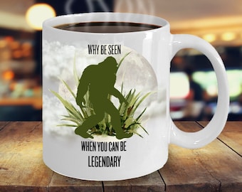 May include: White ceramic coffee mug with a green silhouette of Bigfoot walking in front of a full moon. The text on the mug reads "Why be seen when you can be legendary."