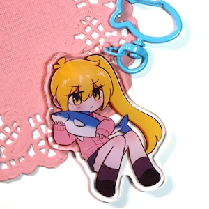 May include: An acrylic keychain featuring an anime-style character with yellow hair in a ponytail, wearing a pink sweater and dark skirt, holding a blue and white shark. The keychain includes a light blue plastic clasp and a fish-shaped ring.