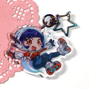 Op de afbeelding: Een doorzichtige acryl sleutelhanger met een cartoonfiguur met donkerblauw haar in een wit en oranje ruimtepak. Het figuur zweeft. De sleutelhanger heeft een stervormige metalen sluiting met een iriserende regenboogafwerking.