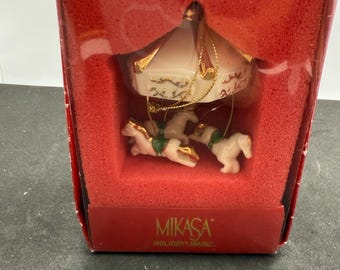 May include: A carousel ornament in a red box. The ornament features a white carousel top with gold accents and three white horses. The box has the text "MIKASA HOLIDAY MAGIC."
