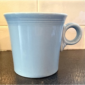 May include: A light blue ceramic mug with a rounded handle. The mug has a cylindrical shape with a slightly flared rim and a series of horizontal ridges near the top. The handle is a loop design.