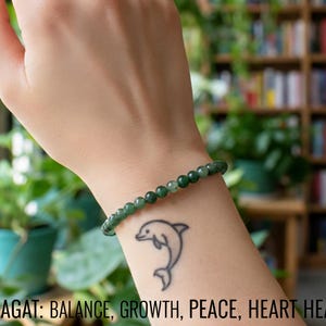 May include: A green moss agate bracelet is worn on a wrist with a dolphin tattoo. The text at the bottom reads "MOSS AGAT: BALANCE, GROWTH, PEACE, HEART HEALING". The background includes plants and bookshelves.