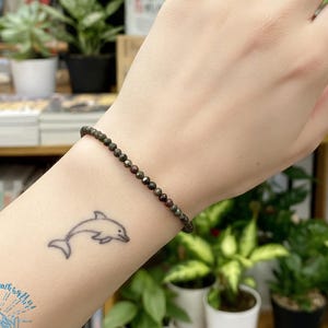 May include: A dark, beaded bracelet with small, round beads is worn on a wrist with a dolphin tattoo. The person's nails are painted a deep red. The background includes plants and bookshelves.