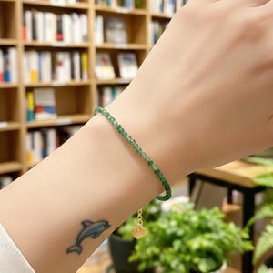May include: A delicate green beaded bracelet with a small gold heart charm. The bracelet is worn on a wrist with a dolphin tattoo. The background includes bookshelves and potted plants, suggesting a natural or bohemian aesthetic.