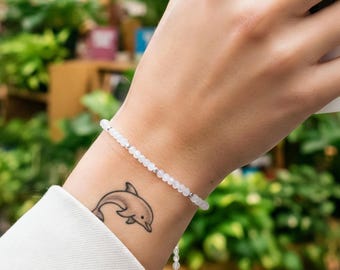 May include: A delicate white beaded bracelet is worn on a wrist with a dolphin tattoo. A person holds a white coffee cup with a brown sleeve. The background features bookshelves and hanging plants.