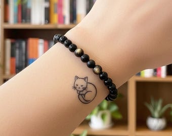 May include: Black beaded bracelet with gold accents. A small cat tattoo is visible on the wrist. The bracelet is made of small, round beads. The background includes a bookshelf with books and plants.