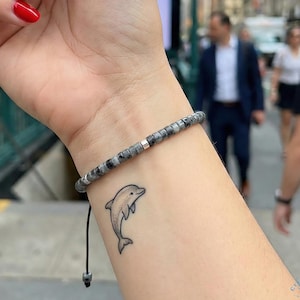 May include: A wrist adorned with a beaded bracelet and a small dolphin tattoo. The bracelet features gray and silver beads. The dolphin tattoo is a simple outline. The person has red nail polish.