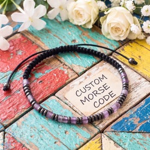 May include: A black cord bracelet featuring alternating metallic dark gray and translucent light purple beads. The bracelet has an adjustable sliding knot closure with two small dark gray beads at the ends. A square card reads CUSTOM MORSE CODE.