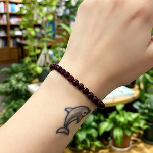 May include: A dark red beaded bracelet is worn on a person's wrist. The bracelet is made of small, round beads. A dolphin tattoo is visible on the person's arm. The background includes plants and bookshelves.