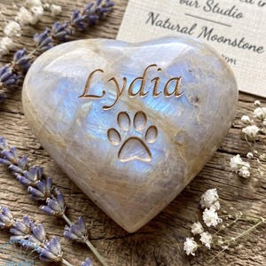 May include: A heart-shaped natural moonstone engraved with the name "Lydia" and a paw print. The stone has a pearlescent sheen with blue and white hues. The text "Hand-engraved in our Studio Natural Moonstone" is on a card.