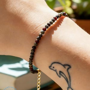 May include: A beaded bracelet with small, round, dark red and green beads. The bracelet is on a wrist with a dolphin tattoo. The bracelet has a gold-colored chain and clasp.