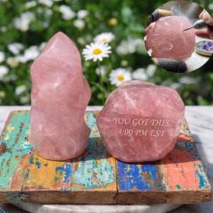 May include: Two pink rose quartz crystals, one a tall, flame-shaped point, the other a rounded shape, sit on a colorful, distressed wooden base. The rounded crystal has the text "YOU GOT THIS. 4:00 PM EST" etched into it. A close-up shows the etching process.