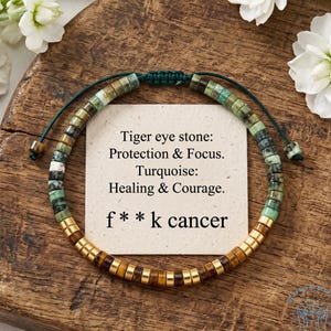 Può includere: Un braccialetto di perline con pietre cilindriche di occhio di tigre e turchese su un cordino verde scuro. Un cartoncino quadrato al centro mostra il testo: Tiger eye stone: Protection & Focus. Turquoise: Healing & Courage. f**k cancer.