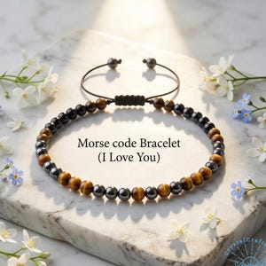 May include: A beaded Morse code bracelet with the message "I Love You". The bracelet features a combination of brown, black, and gold-colored beads. The adjustable cord is black. The bracelet is displayed on a marble surface, surrounded by white and blue flowers.