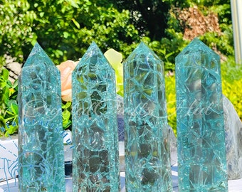 May include: Four turquoise crystal obelisks with a cracked ice effect. The crystals are standing upright, with a pointed top and a rectangular base. The background is blurred, with green foliage and a hint of flowers.