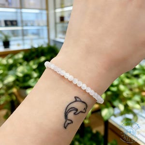 May include: A delicate white beaded bracelet. The bracelet is made of small, round, white beads strung together. The bracelet is worn on a wrist with a black dolphin tattoo. The background is blurred with green foliage.