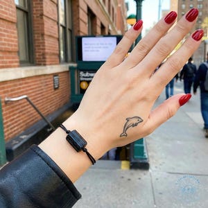 Puede incluir: Una pulsera negra con una piedra negra rectangular se lleva en la muñeca. Un pequeño tatuaje de delfín es visible en la mano. Las uñas están pintadas de rojo. El fondo muestra una calle de la ciudad con una entrada de metro.