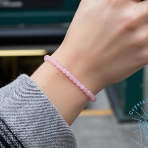Peut inclure: Un bracelet de perles rose clair est porté au poignet. Le bracelet est composé de petites perles rondes. Une tasse à café blanche avec un manchon marron est tenue. L'arrière-plan montre une entrée de métro et une rue de la ville.