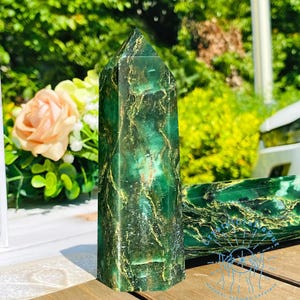 May include: A green and gold crystal point with a marbled pattern. The crystal is on a wooden surface.