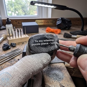 May include: A person using a rotary tool to engrave text on a dark, speckled stone. The text reads "To my missing puzzle piece - Lydia." The workspace includes tools, a lamp, and a sign that says "Crystal Crafts Hut."