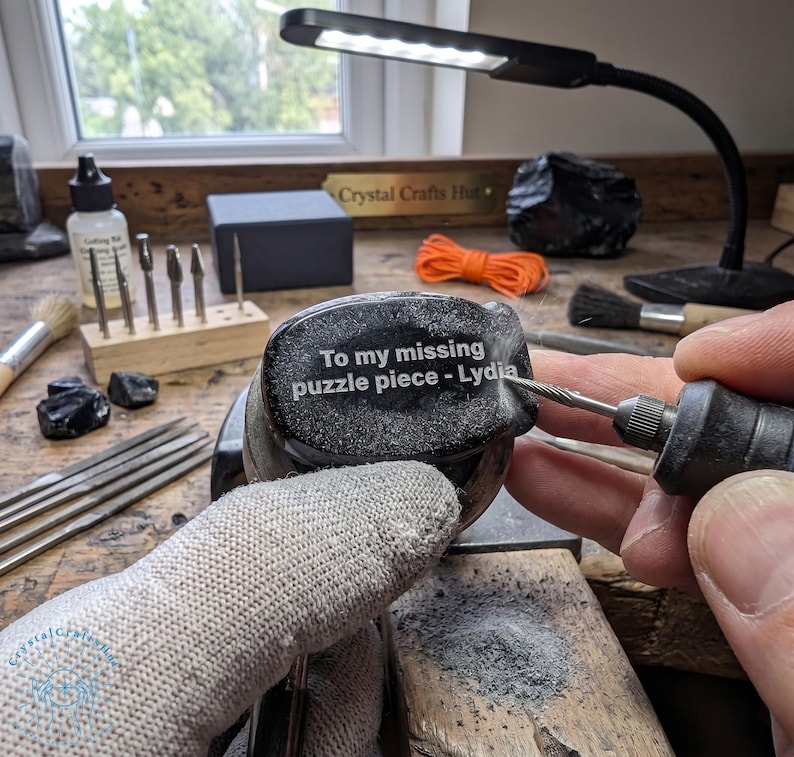 May include: A person using a rotary tool to engrave text on a dark, speckled stone. The text reads "To my missing puzzle piece - Lydia." The workspace includes tools, a lamp, and a sign that says "Crystal Crafts Hut."
