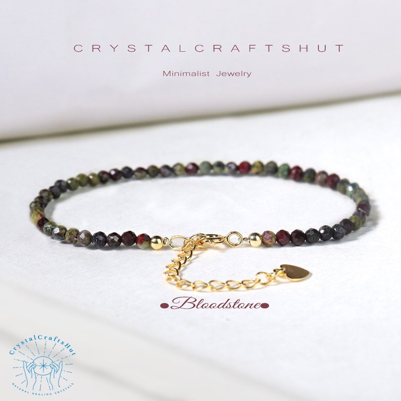 May include: A minimalist bracelet featuring faceted bloodstone beads in shades of green and burgundy. The bracelet has a gold-colored chain and clasp, with a small heart-shaped charm. The text "CRYSTALCRAFTSHUT" and "Minimalist Jewelry" are visible.