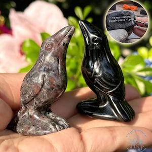 May include: Two carved stone raven figurines. One is a speckled gray and brown, the other is solid black. The ravens are perched on a hand. A small inset image shows a stone with the text "To my missing puzzle piece - Lydia."