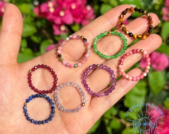 Peut inclure: Plusieurs bagues composées de perles de différentes couleurs : rouge, violet, vert, rose et bleu. Chaque bague est ornée de petites perles rondes et d'un détail doré. Les bagues sont présentées sur une main, avec un fond de feuillage vert et de fleurs roses.