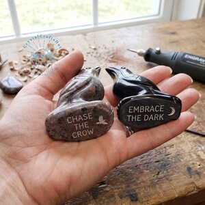 May include: Two carved stone raven figurines. One is a speckled brown color, and the other is black. The brown raven has the text "CHASE THE CROW" and a bird graphic. The black raven has the text "EMBRACE THE DARK" and a crescent moon graphic.
