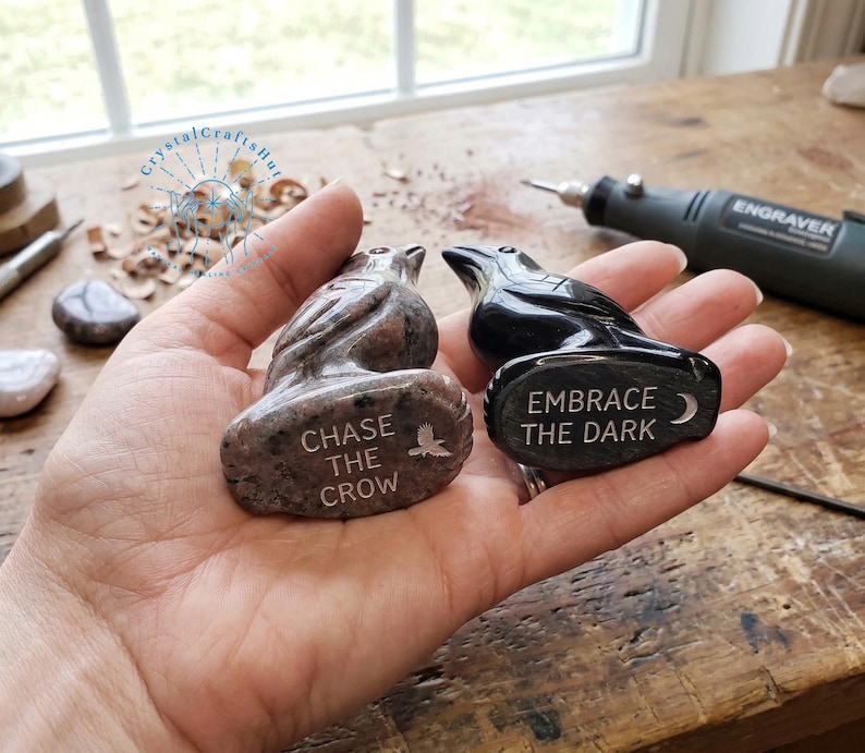 May include: Two carved stone raven figurines. One is a speckled brown color, and the other is black. The brown raven has the text "CHASE THE CROW" and a bird graphic. The black raven has the text "EMBRACE THE DARK" and a crescent moon graphic.