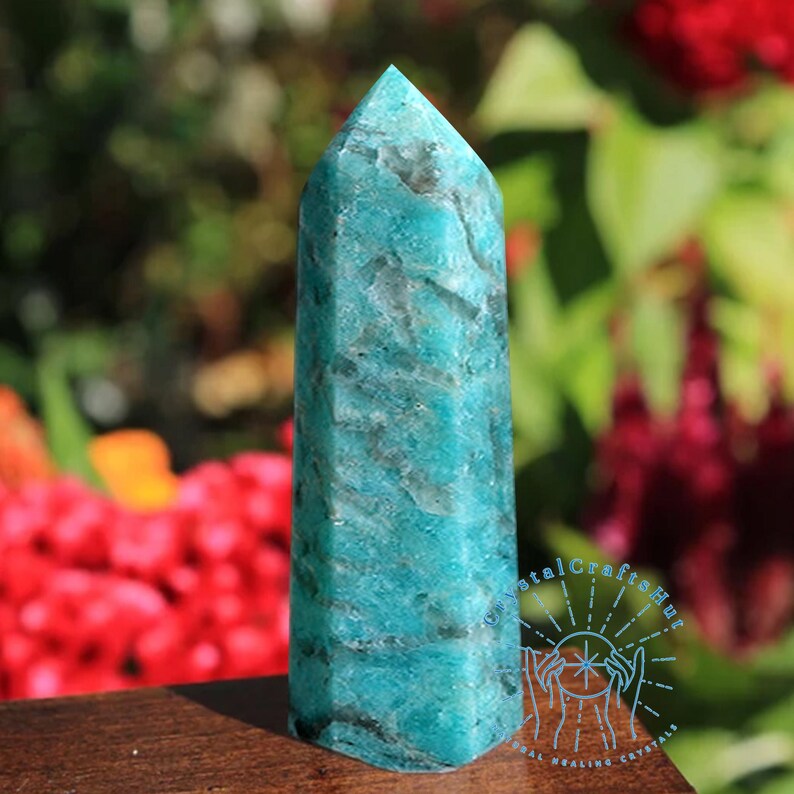 Puede incluir: Un obelisco de cristal de amazonita turquesa con una parte superior puntiaguda. La piedra tiene un aspecto veteado con vetas de color gris oscuro. El obelisco se exhibe sobre una superficie de madera, con un fondo borroso de follaje rojo y verde.