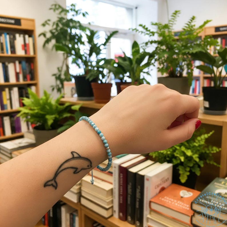 May include: A light blue beaded bracelet with a silver accent is worn on a person's wrist. A black and white dolphin tattoo is visible on the forearm. The background includes bookshelves and potted plants.