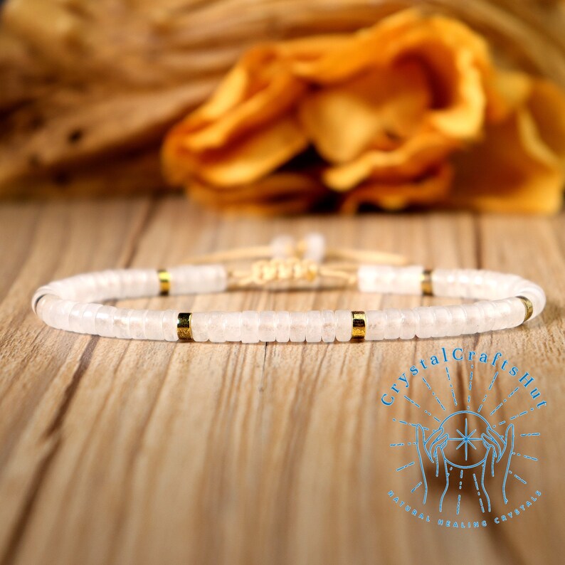 May include: A white beaded bracelet with gold-colored accents. The bracelet is made of small, flat, round beads and has three gold-colored spacers. The bracelet is on a wooden surface.
