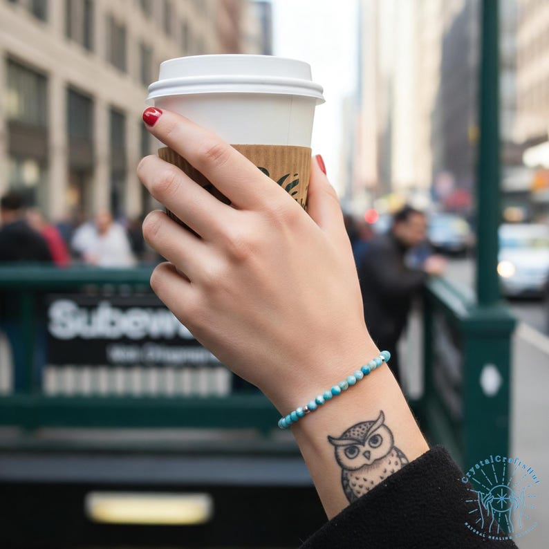 May include: A person holding a white paper coffee cup with a brown sleeve, wearing a blue and silver beaded bracelet. An owl tattoo is visible on the wrist. The background shows a city street and a subway entrance.