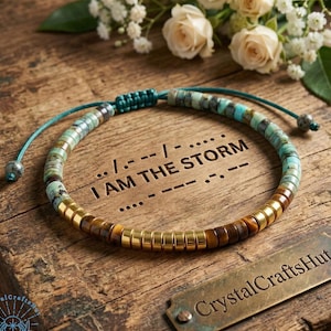 May include: A beaded bracelet with turquoise, brown, and gold-colored beads. The bracelet is on a wooden surface with the text "I AM THE STORM" and Morse code. A brass tag reads "CrystalCrafts Hut".