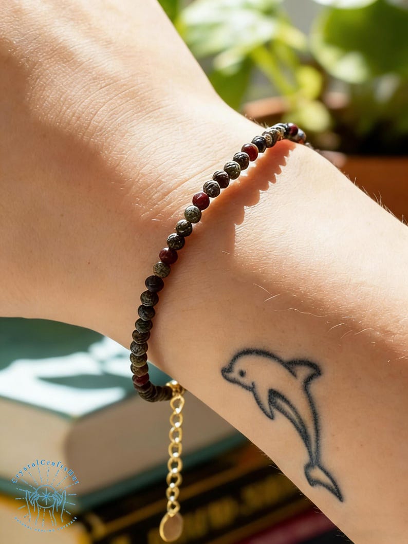 May include: A beaded bracelet with small, round, dark red and green beads. The bracelet is on a wrist with a dolphin tattoo. The bracelet has a gold-colored chain and clasp.