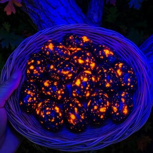 May include: A woven basket filled with glowing, egg-shaped stones. The stones are dark blue with vibrant orange and yellow speckles, creating a striking contrast. The basket is held by a hand, with a dark background of foliage.
