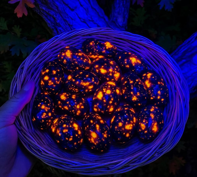 May include: A woven basket filled with glowing, egg-shaped stones. The stones are dark blue with vibrant orange and yellow speckles, creating a striking contrast. The basket is held by a hand, with a dark background of foliage.