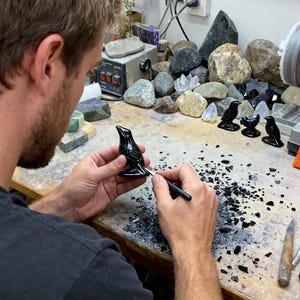 May include: A person working on a black bird figurine with a tool. Several other bird figurines are on the table, along with an orange bag that says "crystalcrafthel". A grinding wheel and various stones are in the background.